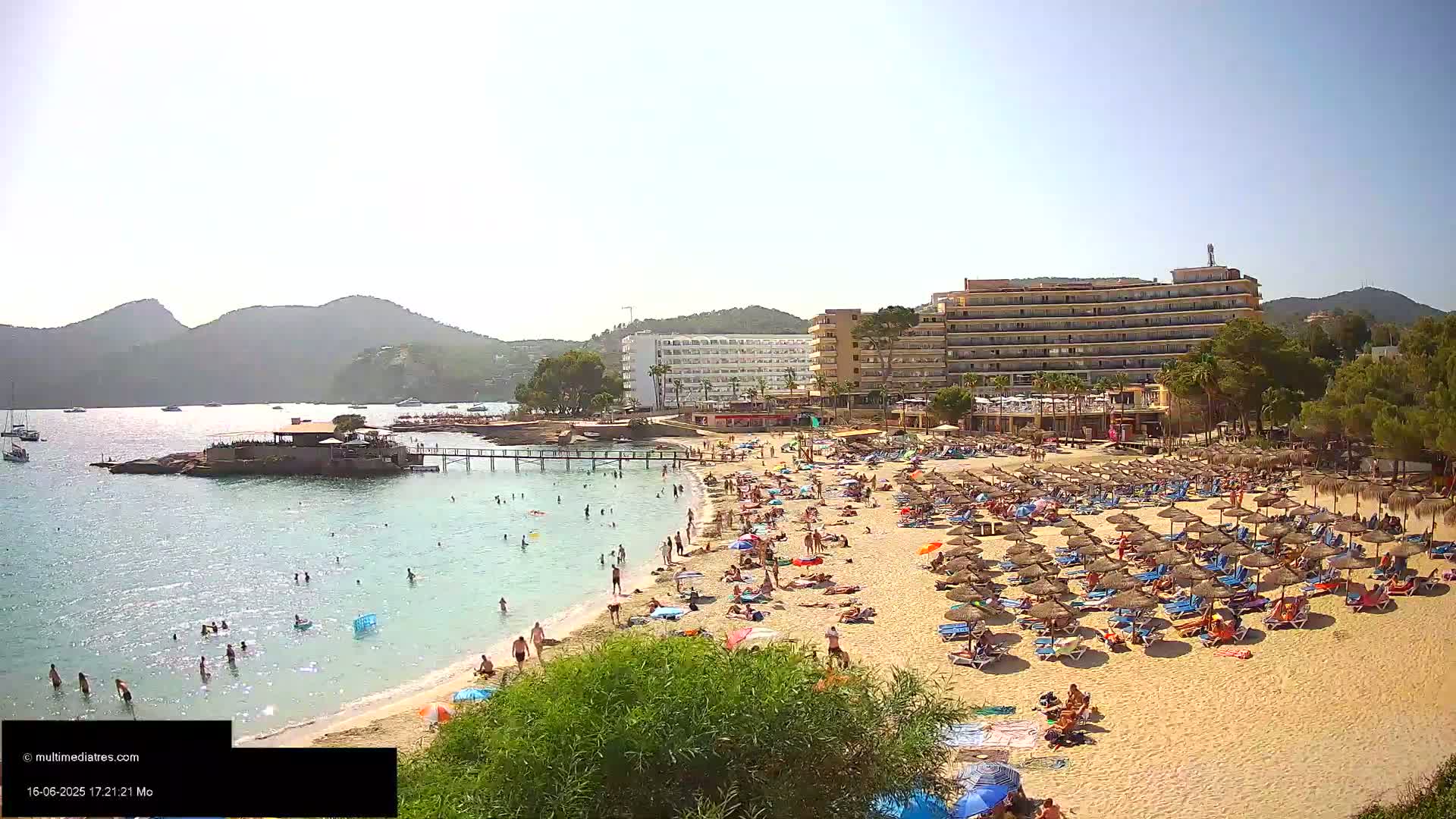 A sandy beach with many people and umbrellas is situated in front of several hotels and a calm bay, under a mostly sunny sky.