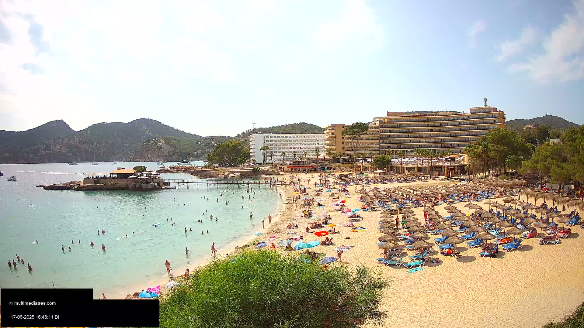 A sandy beach with numerous umbrellas and lounge chairs is crowded with people, backed by hotels and green hills under a bright, sunny sky.