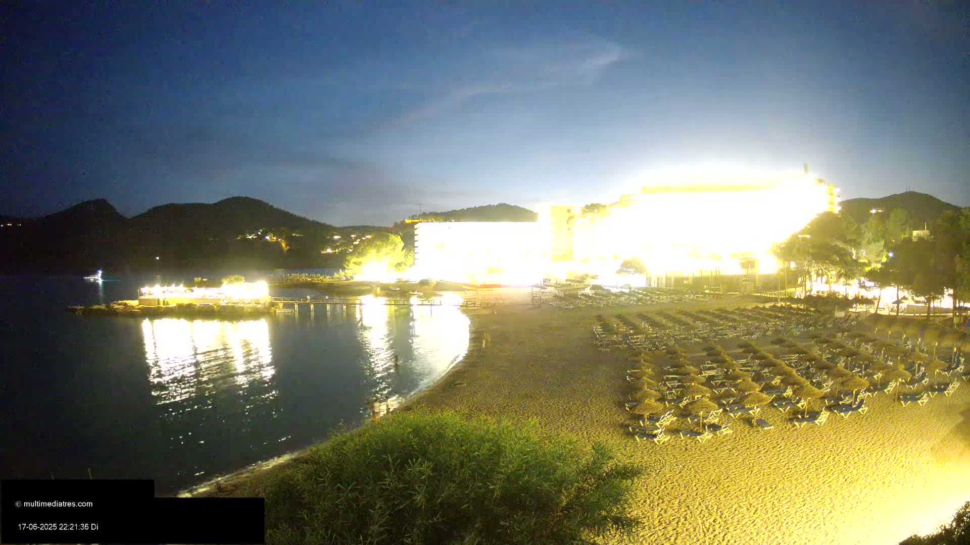 A brightly lit beach at night, with a large hotel behind it,  is shown under a dark, clear sky.