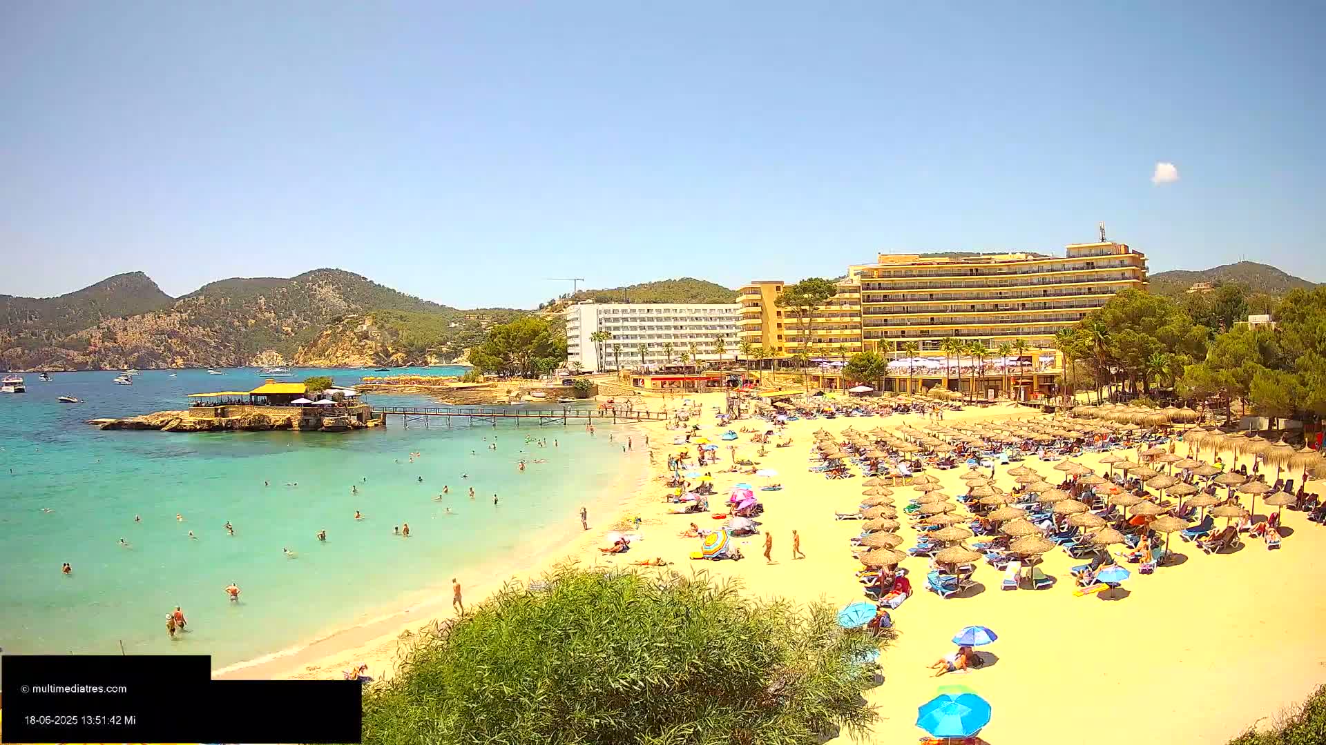 A sunny beach scene shows a sandy shore crowded with beach umbrellas and people, turquoise water, a nearby restaurant on a small island, and hotels behind the beach.