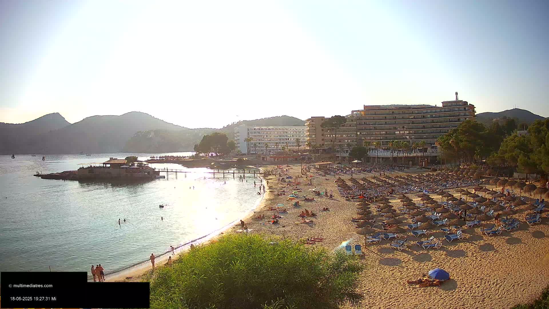 A sandy beach with numerous beach umbrellas and sunbathers is situated next to calm, clear water, backed by hotels and hills under a bright, sunny sky.