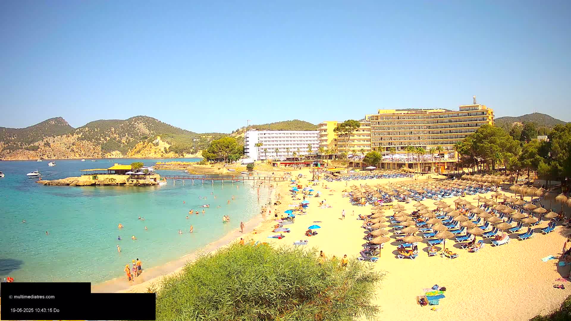 A sunny beach scene shows a sandy shore lined with thatched umbrellas and lounge chairs, many people swimming and relaxing, a hotel in the background, and a small island with a restaurant in the turquoise water.