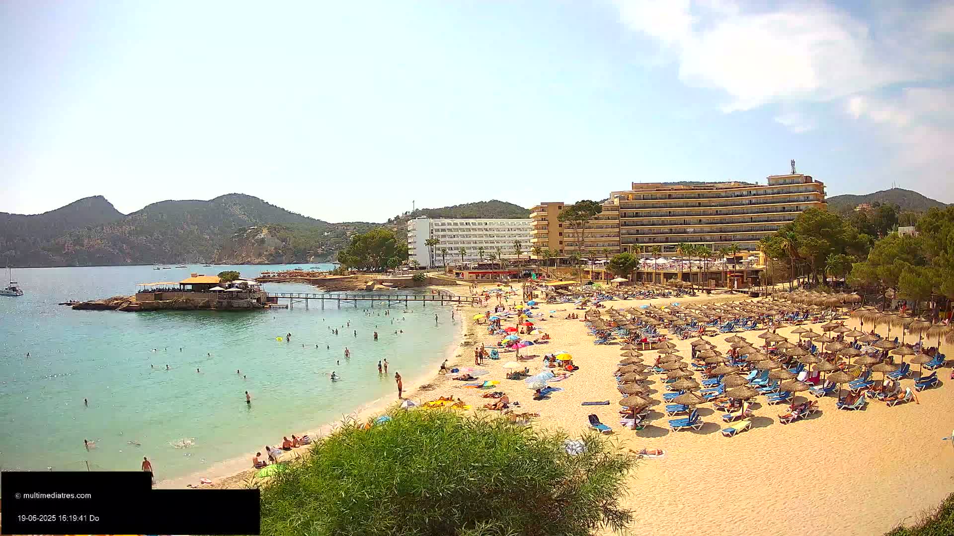 A sunny beach scene shows numerous people relaxing under umbrellas on a sandy beach near hotels and a calm, clear body of water with people swimming.
