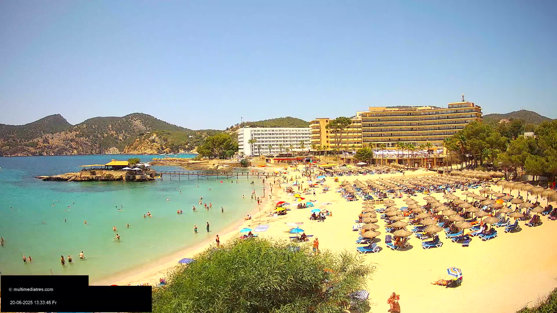 A sunny beach scene shows a sandy shore lined with numerous thatched umbrellas and beach chairs, with people swimming and sunbathing in the turquoise water, backed by hotels and lush green hills.