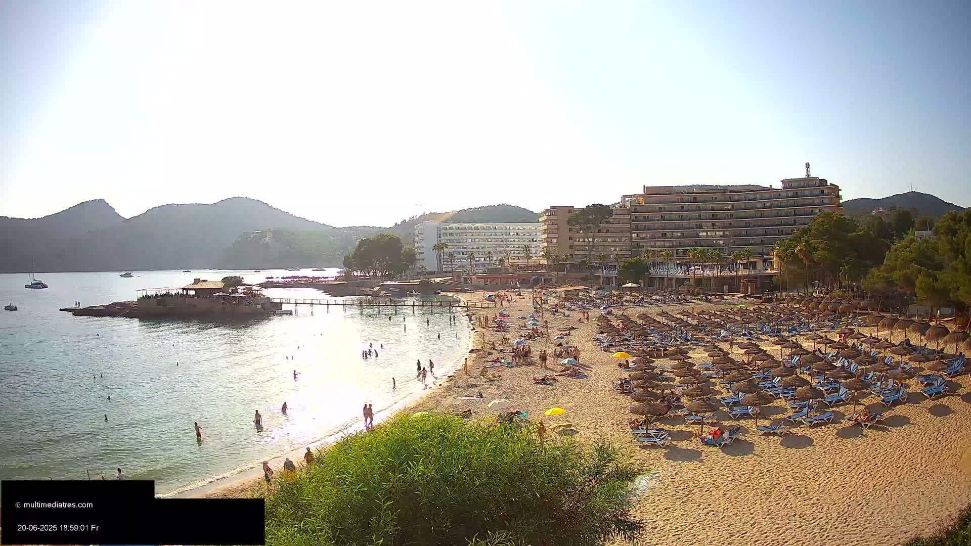 A sandy beach with numerous umbrellas and lounge chairs is situated below a hotel and other buildings,  with people swimming and relaxing in the calm, sunny water and mountains in the background.