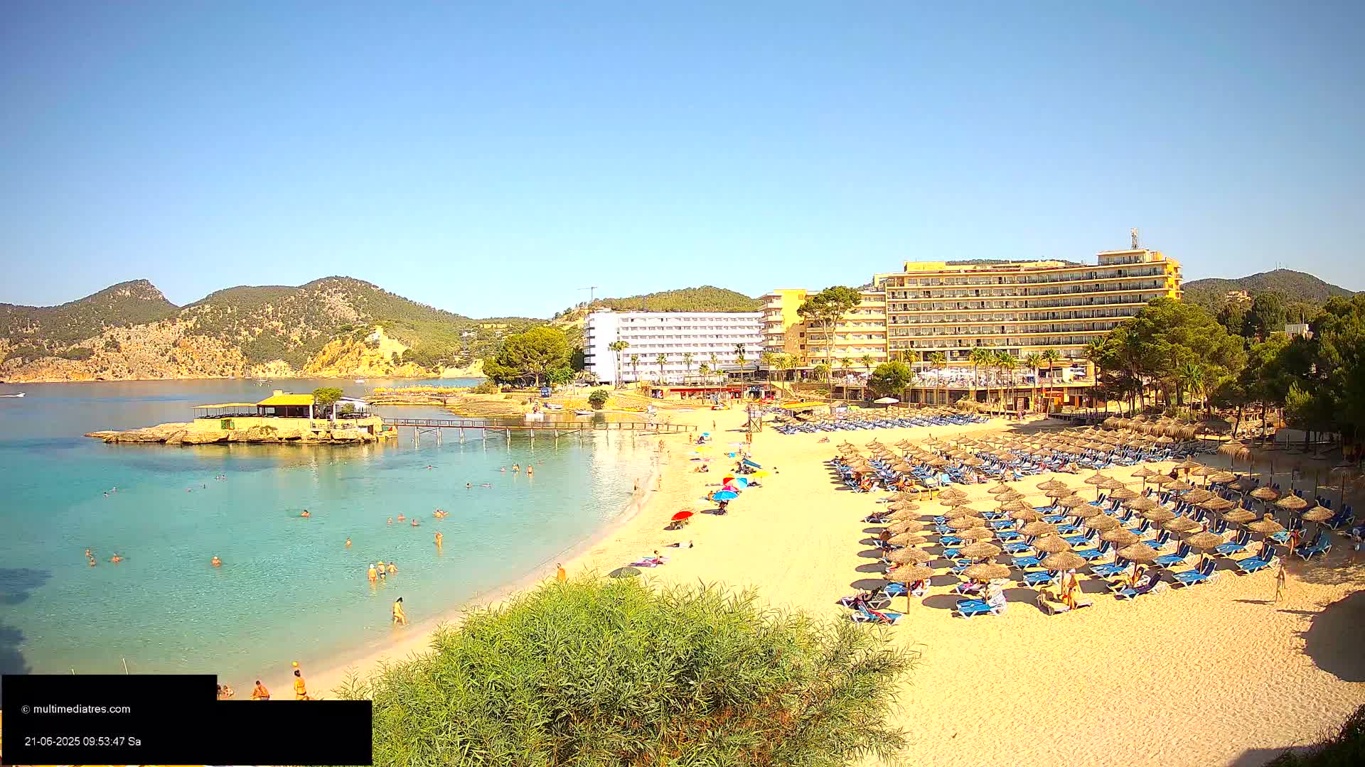 A sandy beach with numerous umbrellas and lounge chairs sits in front of a large hotel, with people swimming and sunbathing in the calm, clear water on a sunny day.