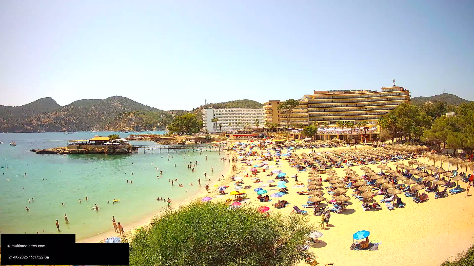 A crowded sandy beach with numerous umbrellas and sunbathers, adjacent to a calm, clear turquoise sea, with hotels and greenery behind, all under a sunny sky.