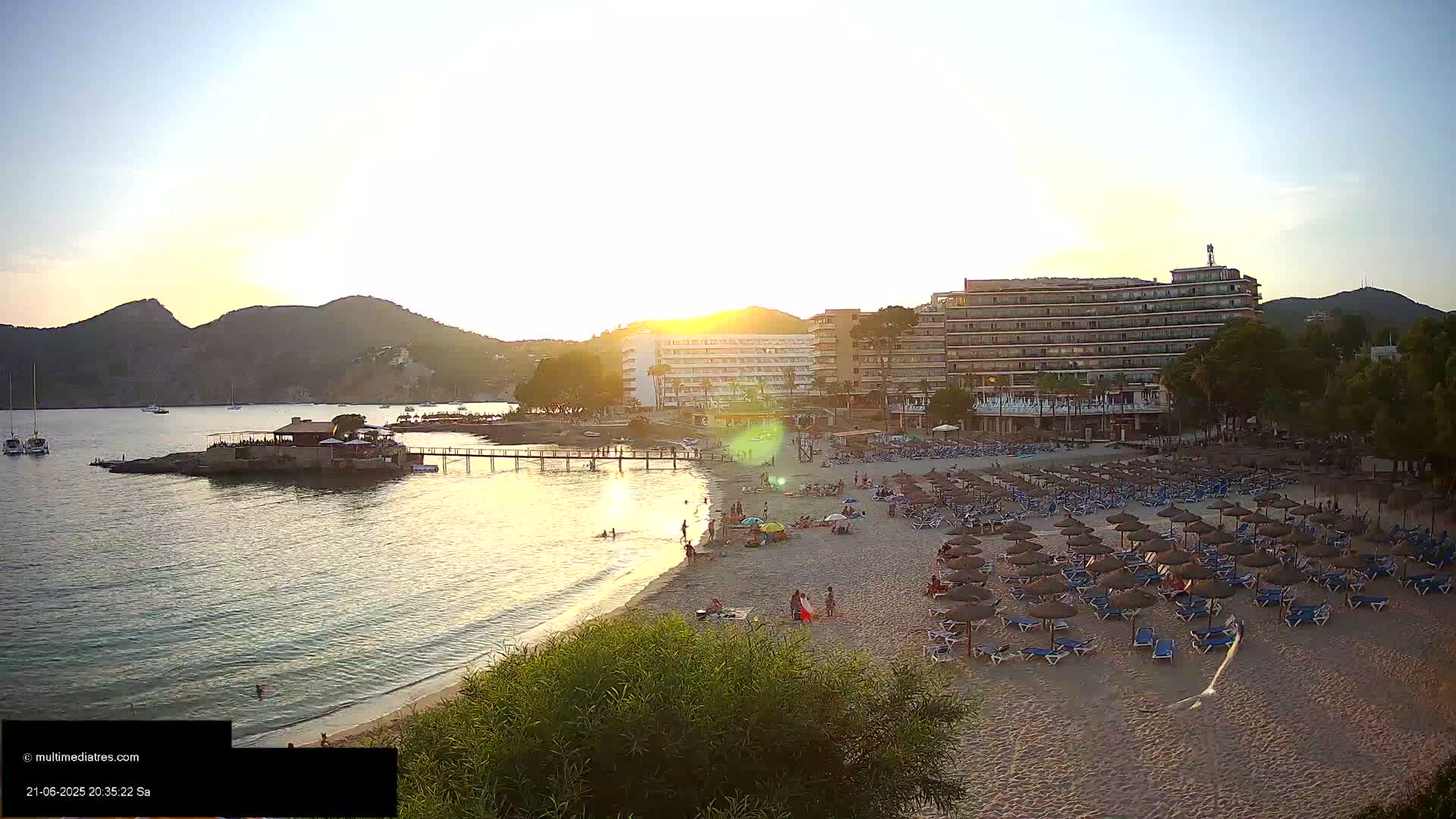 A sandy beach with numerous umbrellas and lounge chairs is situated next to a large hotel at sunset, with calm water and boats visible in the distance.