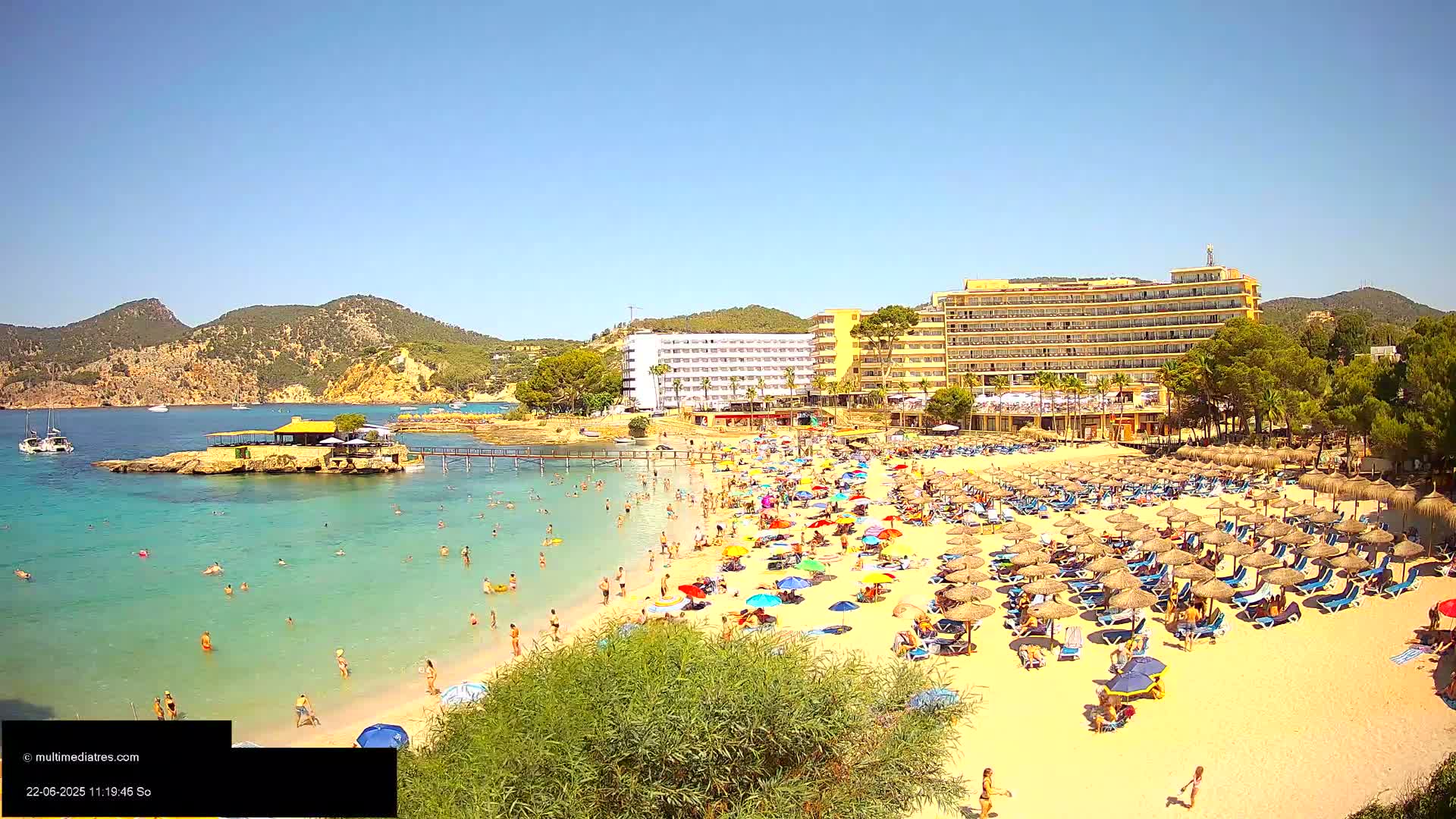 A sunny beach scene shows a sandy shore crowded with people under numerous umbrellas, adjacent to a large hotel and calm, turquoise water.