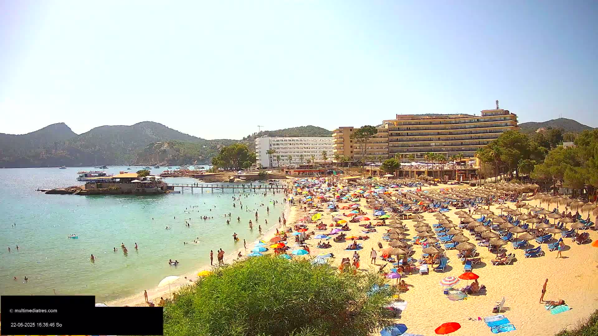 A crowded sandy beach with numerous colorful umbrellas and lounge chairs, adjacent to a calm, clear bay with swimmers, and backed by hotels and lush green hills under a sunny sky.