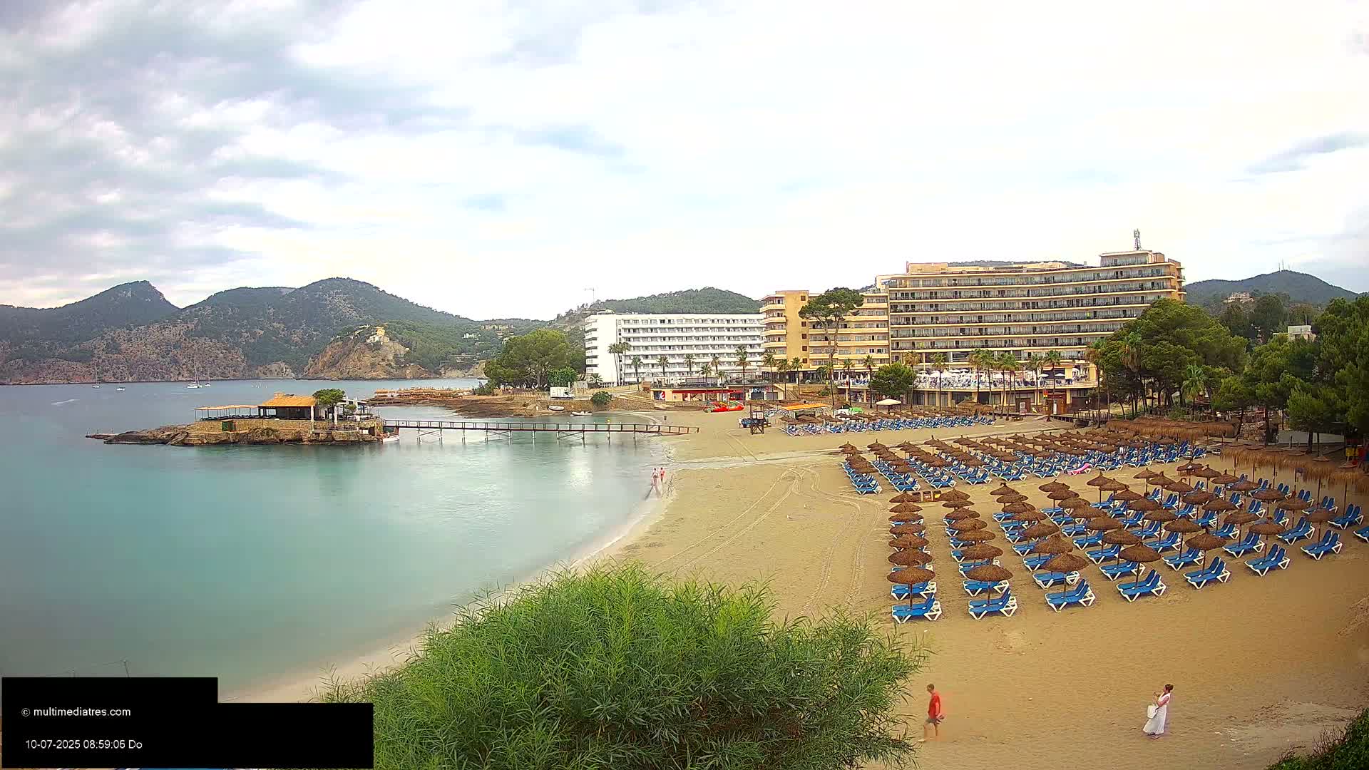 A sandy beach with numerous blue lounge chairs and thatched umbrellas is situated in front of several hotels, overlooking a calm bay and mountains under a cloudy sky.