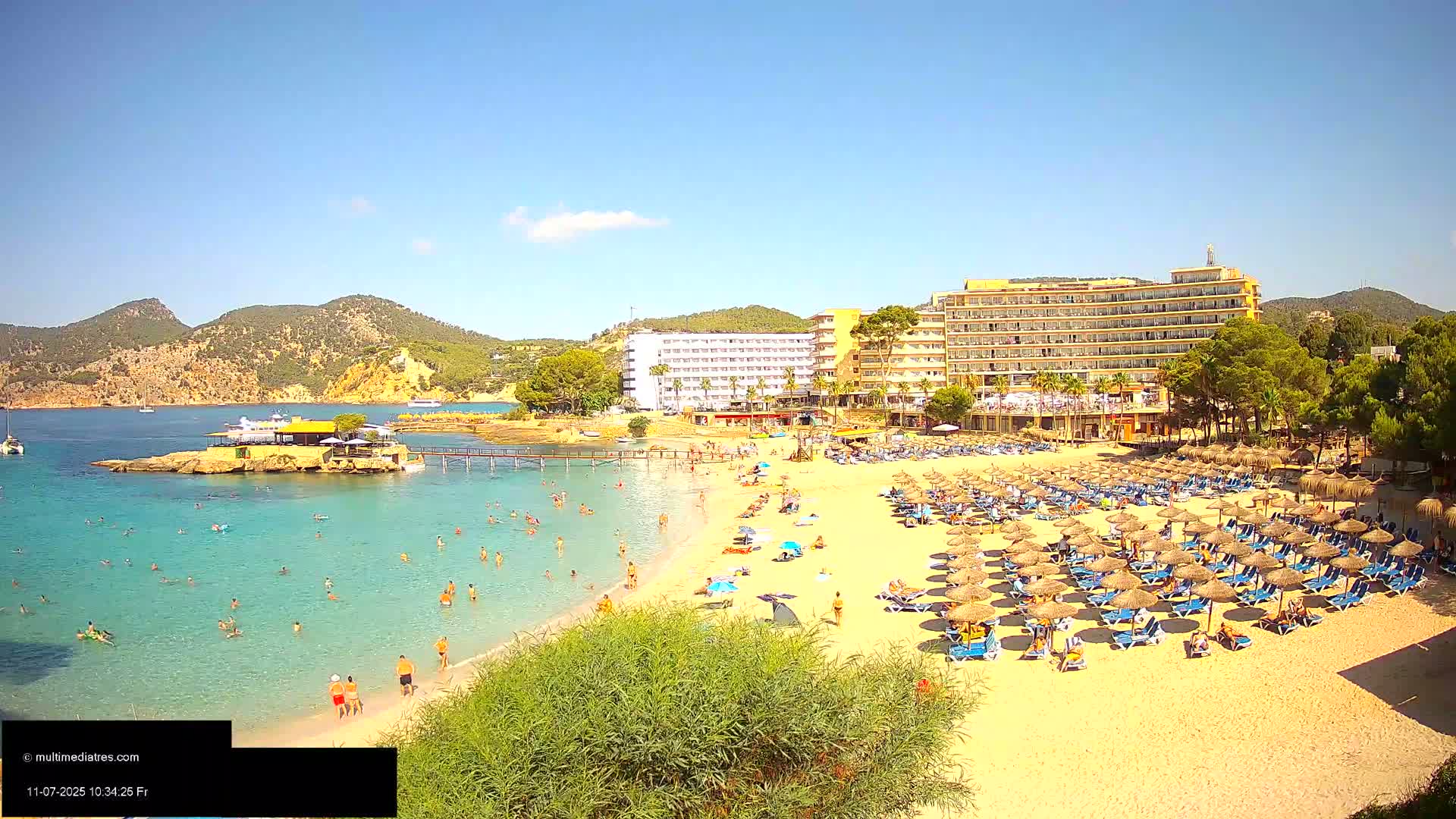 A sandy beach with numerous umbrellas and sunbathers is adjacent to a large hotel, all situated on a clear, turquoise bay under a sunny sky.