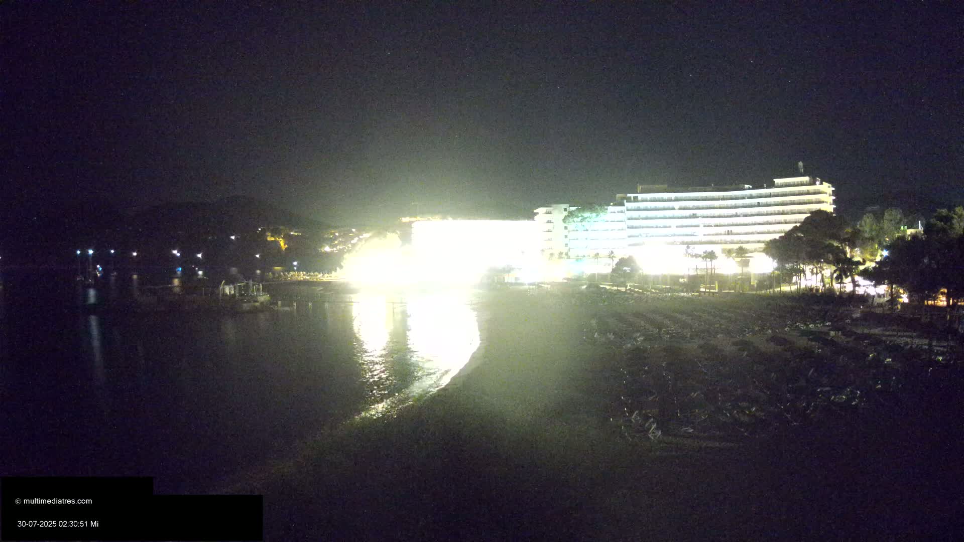 A sandy beach with numerous umbrellas and lounge chairs sits below two large hotels near a calm, clear body of water under a partly cloudy sky.