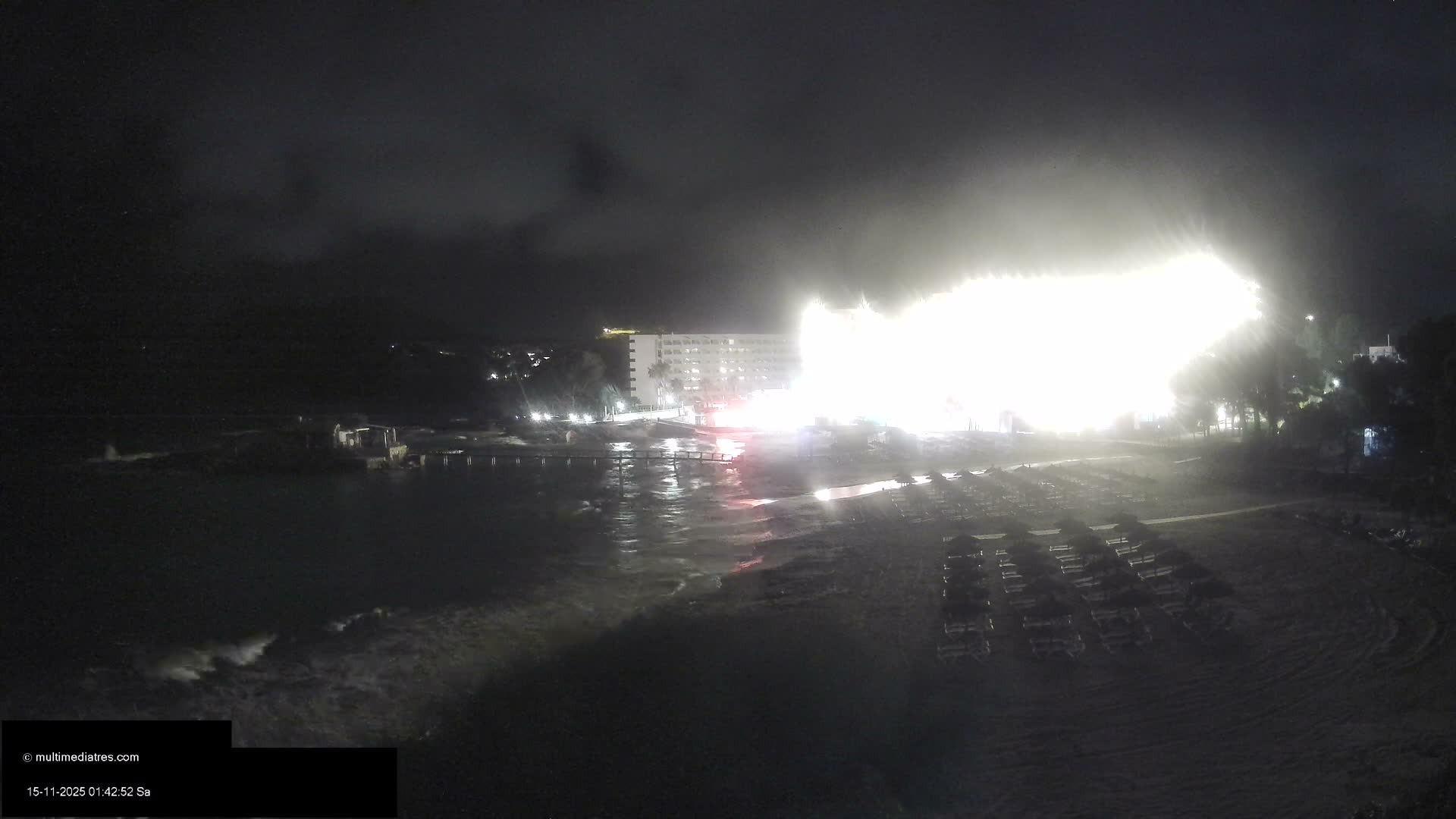 An expansive beach and resort complex are illuminated by an extremely bright, almost blinding light source at night, with a multi-story hotel and rows of sun loungers visible under an overcast sky.