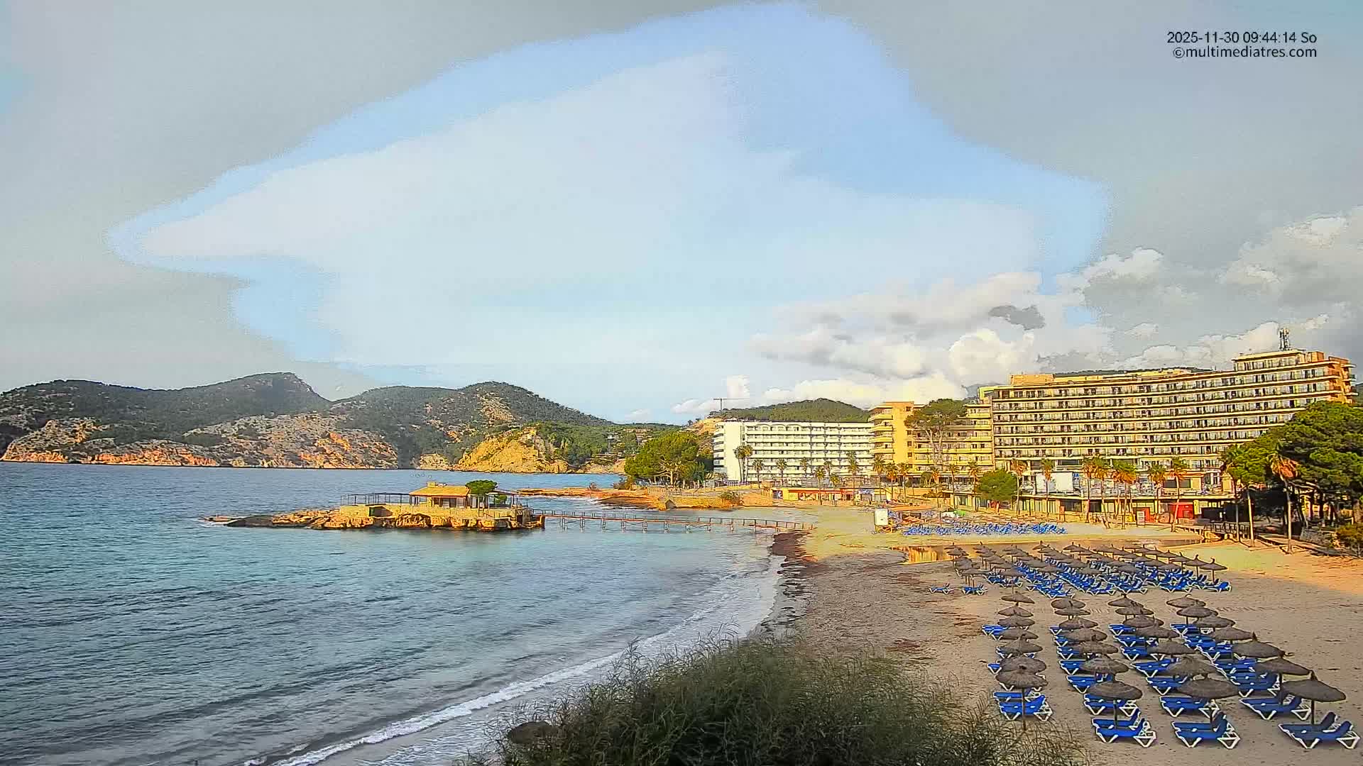The image displays a large beach bay lined with resort hotels, featuring rows of unoccupied beach chairs and straw umbrellas on the sandy shore, a building on a rocky islet connected by a pier, and mountains in the distance, all under a partially cloudy sky.