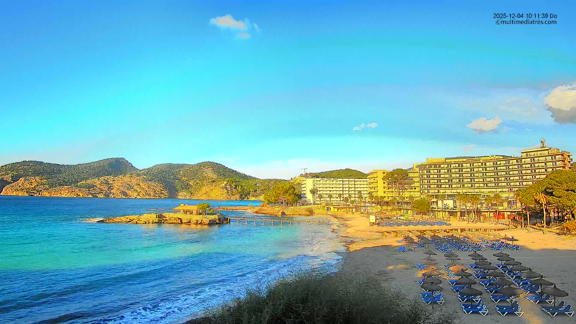 The image shows a sunny coastal scene with a sandy beach lined by hotels, rows of sun loungers and straw umbrellas, facing clear turquoise waters with a small pier and rocky islets, all set against a backdrop of green mountains under a bright blue sky with scattered clouds.