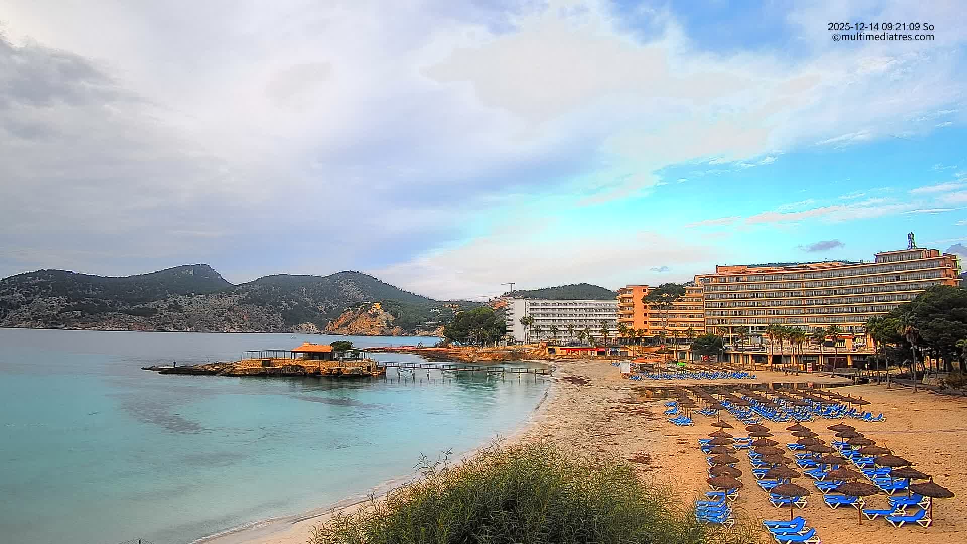 The image shows a sunny coastal scene with a sandy beach lined by hotels, rows of sun loungers and straw umbrellas, facing clear turquoise waters with a small pier and rocky islets, all set against a backdrop of green mountains under a bright blue sky with scattered clouds.