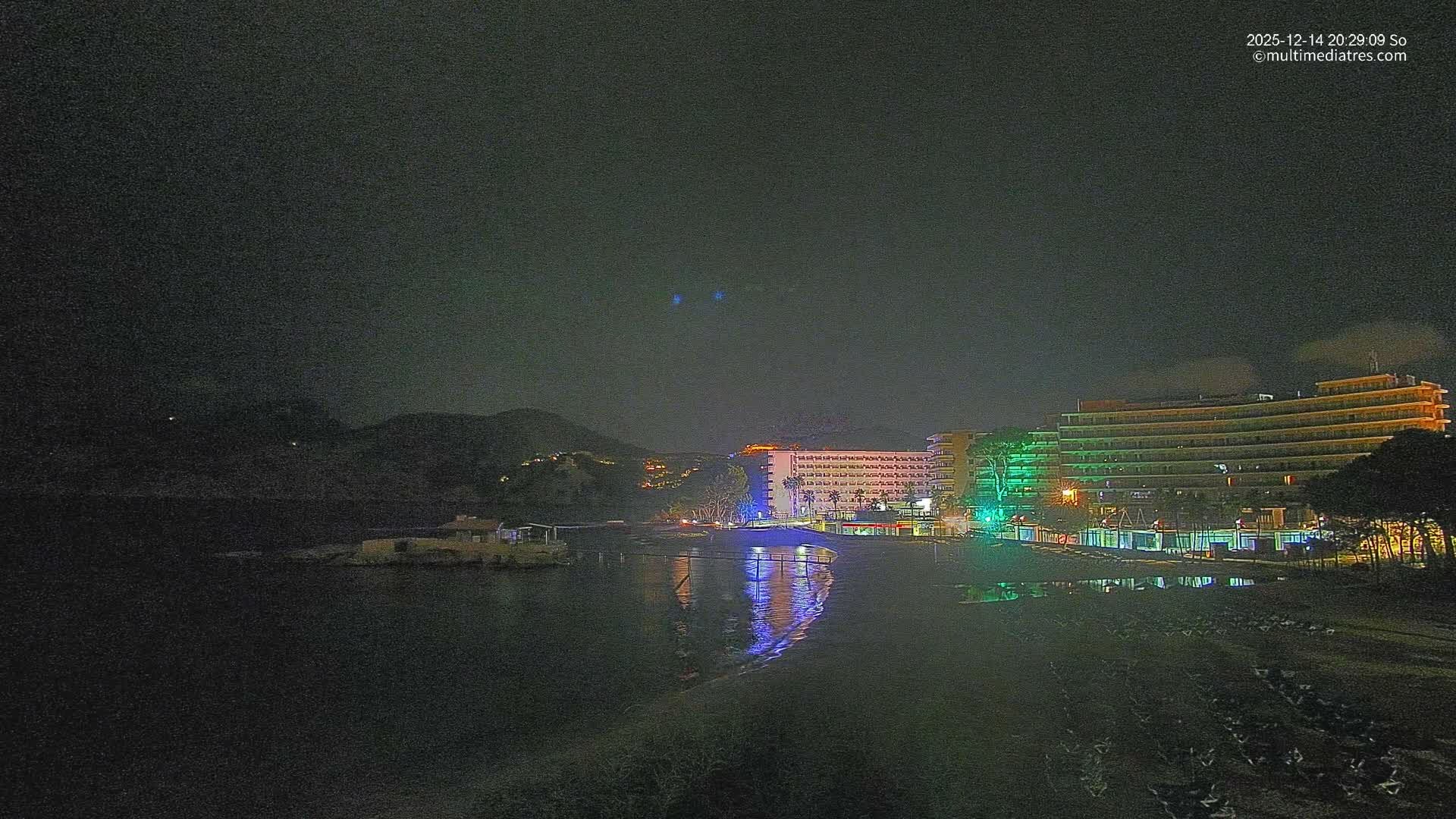 The image shows a sunny coastal scene with a sandy beach lined by hotels, rows of sun loungers and straw umbrellas, facing clear turquoise waters with a small pier and rocky islets, all set against a backdrop of green mountains under a bright blue sky with scattered clouds.