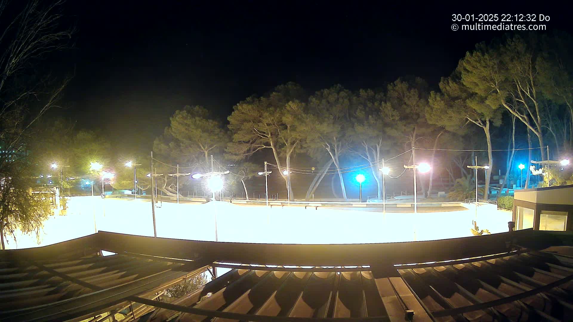 A brightly lit, empty outdoor plaza at night, surrounded by tall trees under a clear, dark sky.
