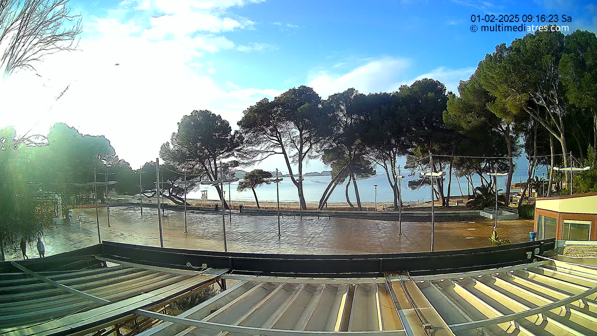 A wet paved plaza overlooks a calm, blue sea and sandy beach lined with pine trees under a partly cloudy sky.