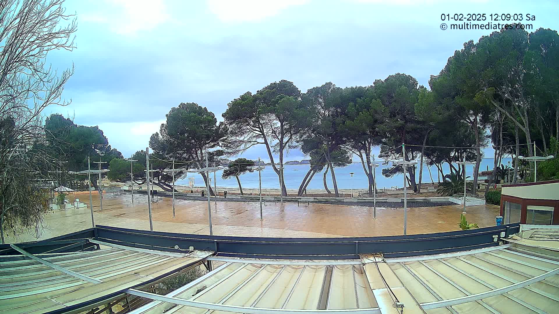A mostly cloudy day reveals a paved plaza overlooking a calm, blue ocean beach lined with pine trees.