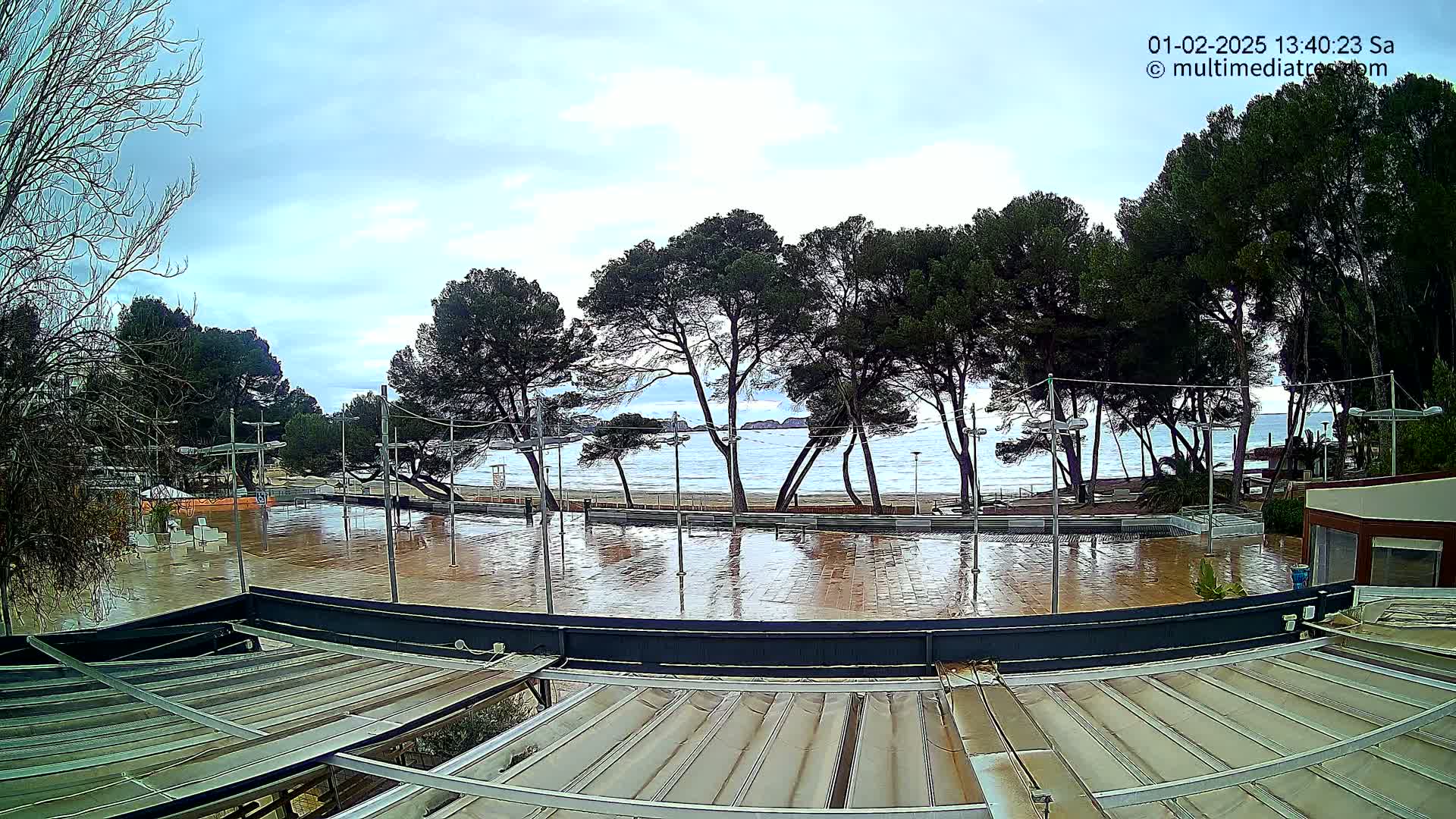 A flooded plaza next to a beach with overcast skies and a calm sea is seen from above a slatted awning.