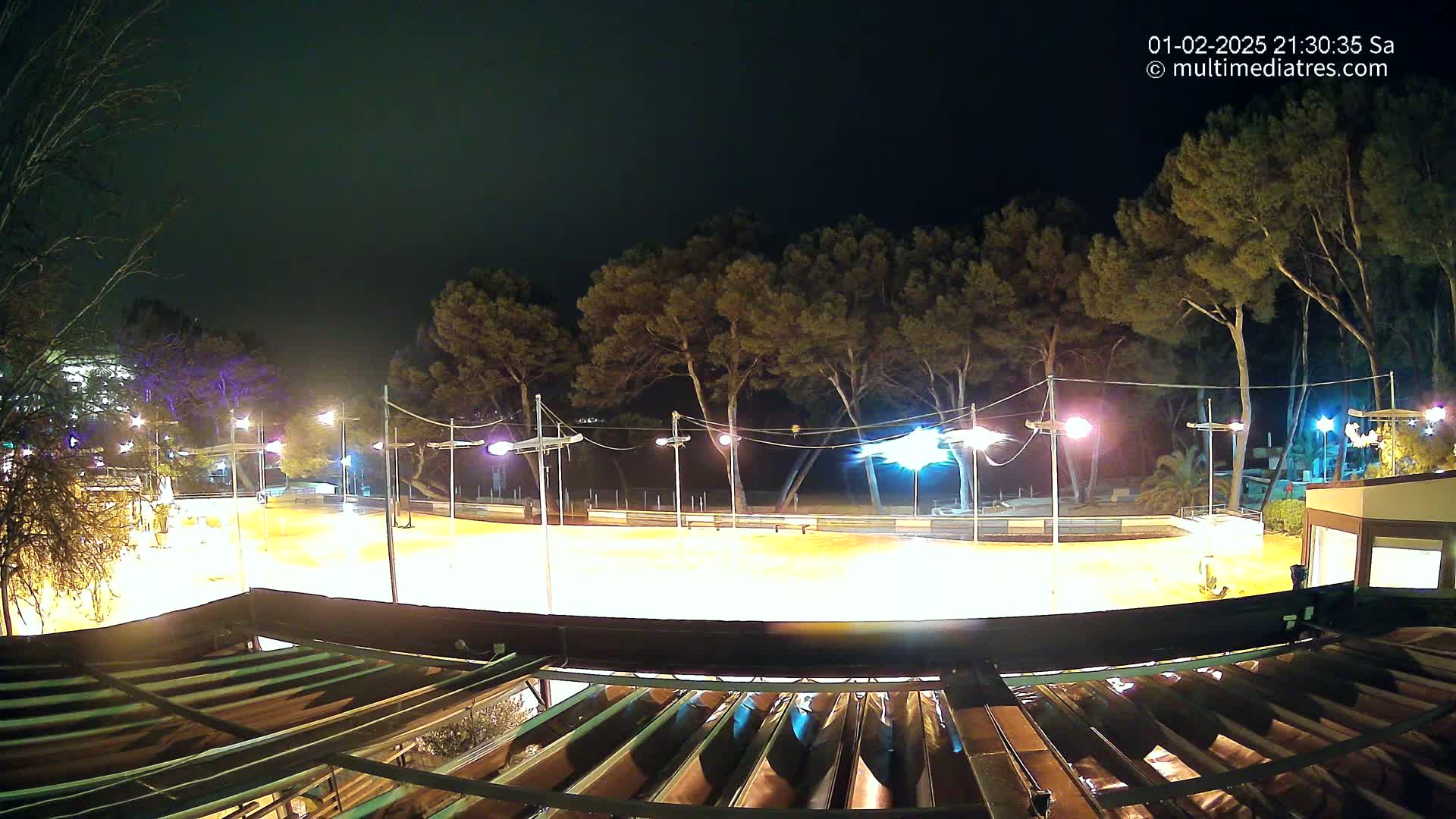 A brightly lit outdoor rink at night, surrounded by trees and partially visible buildings under a dark, clear sky.