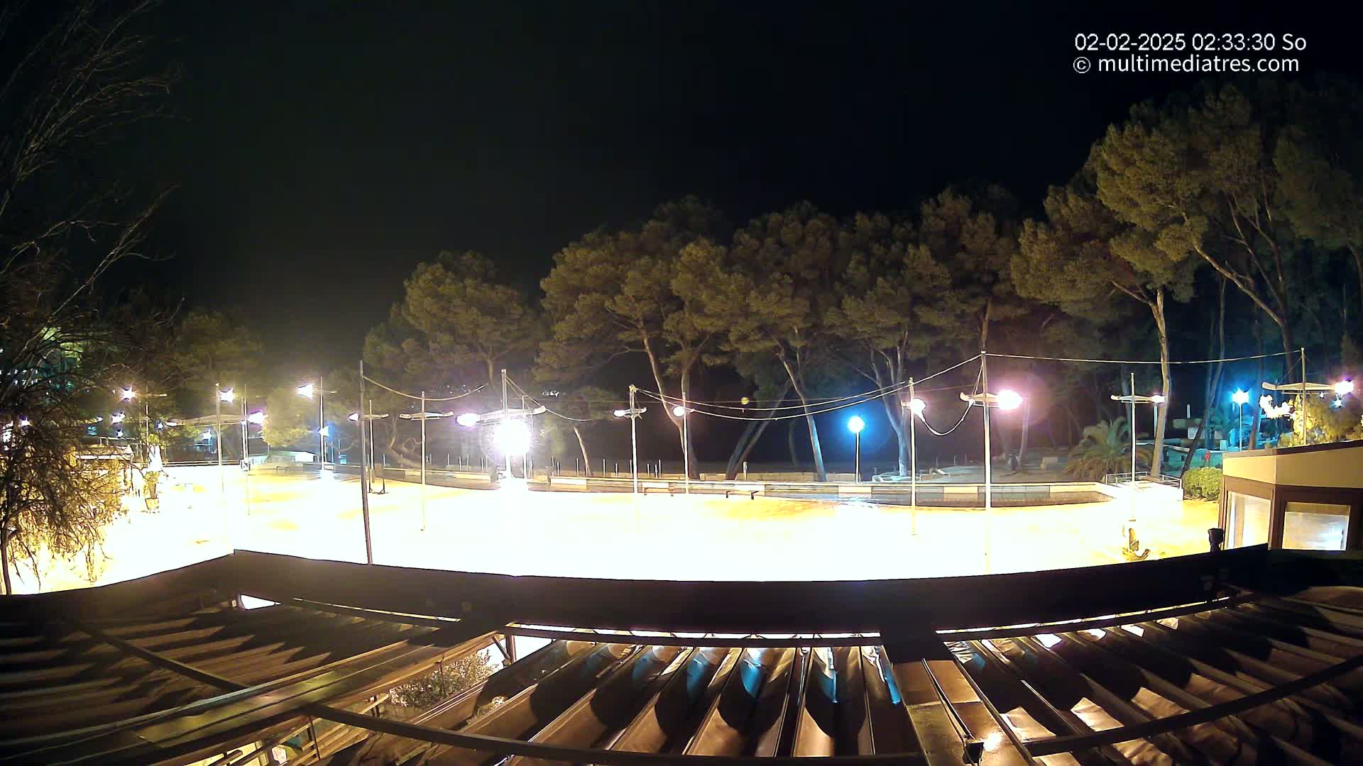 A brightly lit, empty outdoor area, possibly a plaza or court, is surrounded by trees at night.