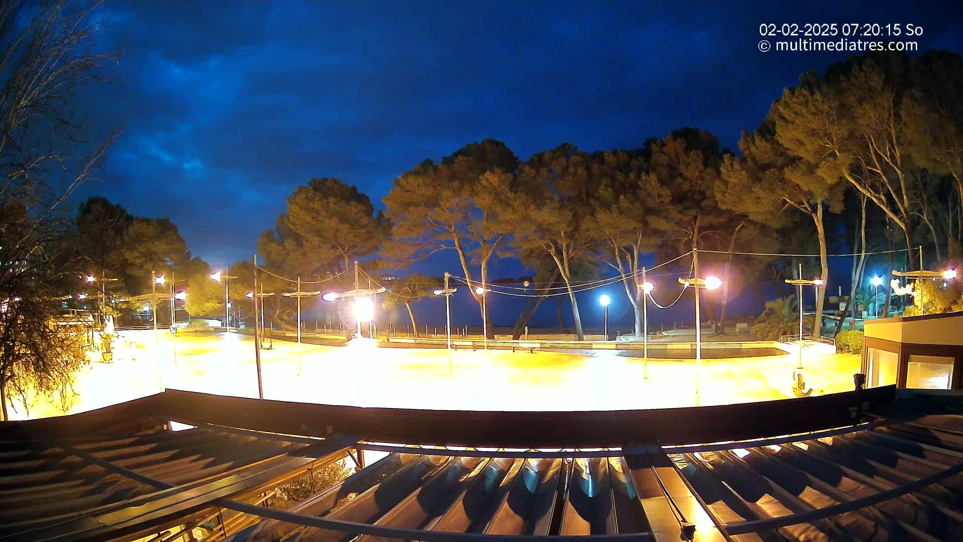 A brightly lit, empty plaza at night, bordered by trees and a calm body of water under a twilight sky.
