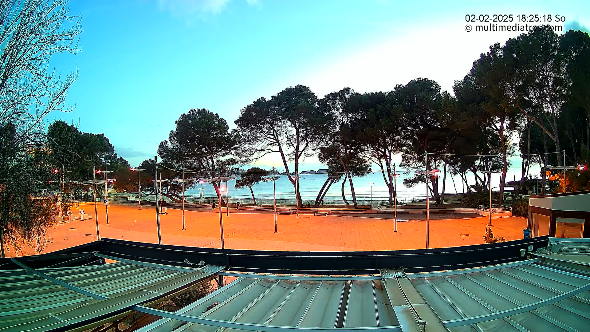 An empty, brightly lit plaza overlooks a calm sea at dusk, with a line of pine trees separating the two.