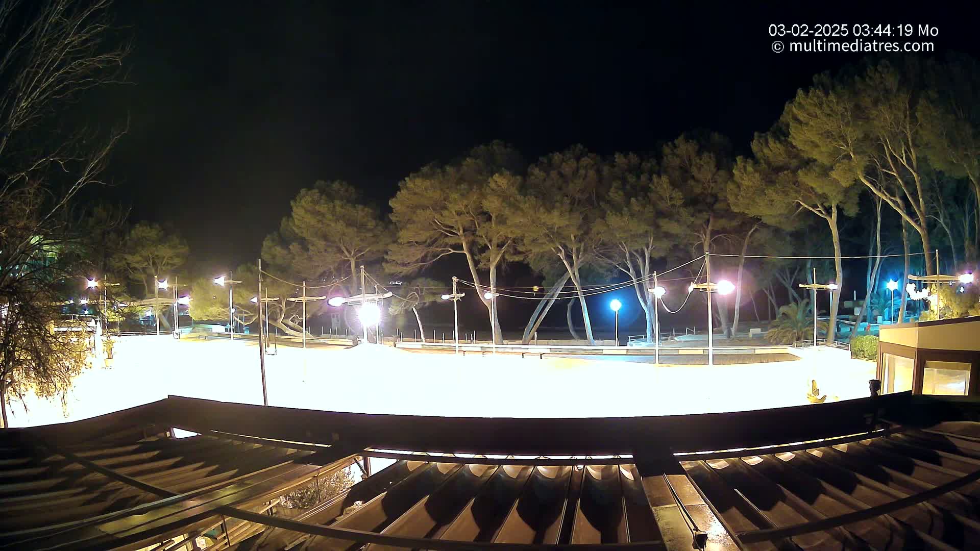 A brightly lit, empty plaza at night, surrounded by tall pine trees under a clear, dark sky, is viewed from above a partially covered patio.