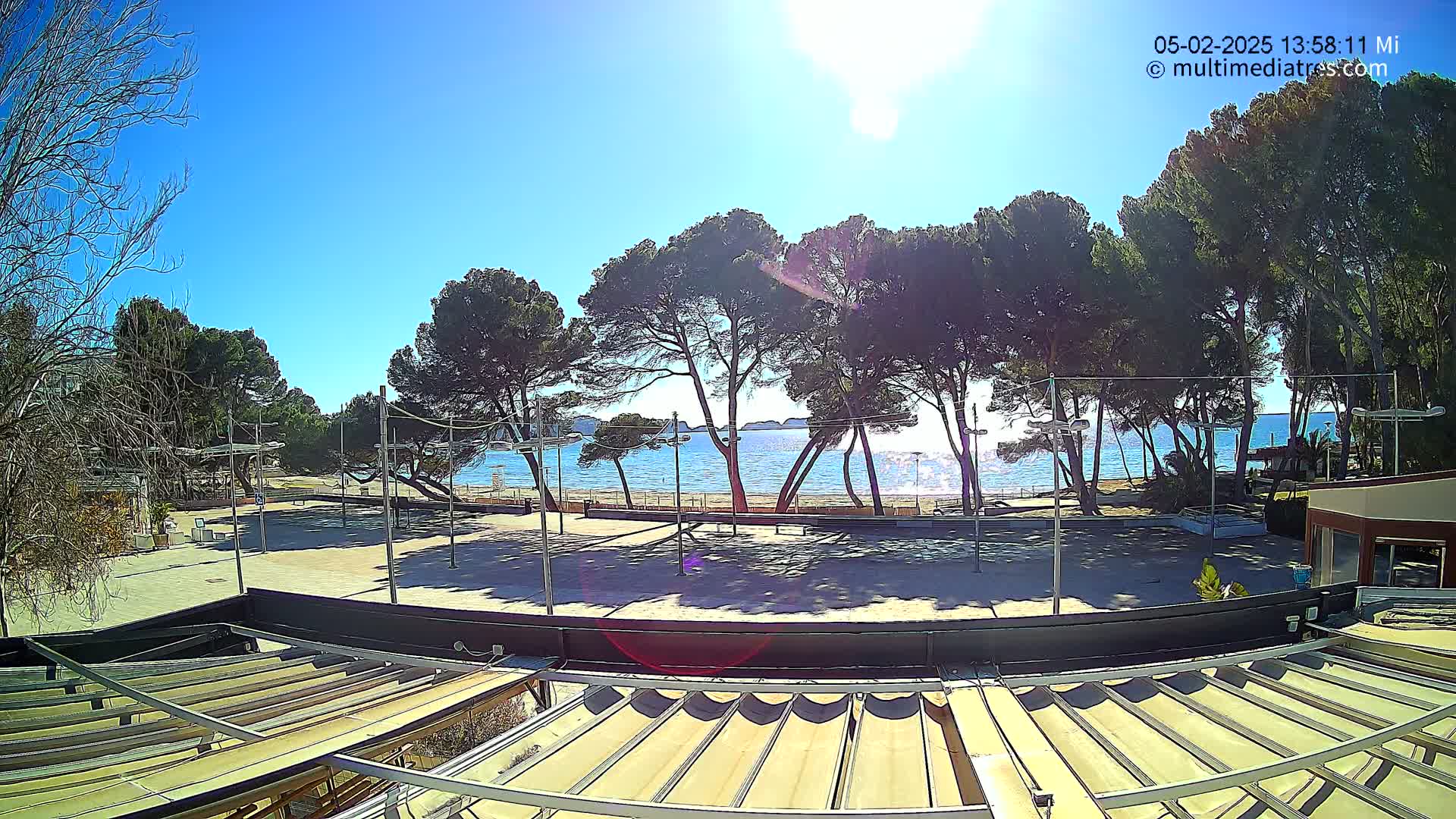 A sunny day at a beachside location shows a sandy area with volleyball courts and several pine trees, beyond which is a calm sea.