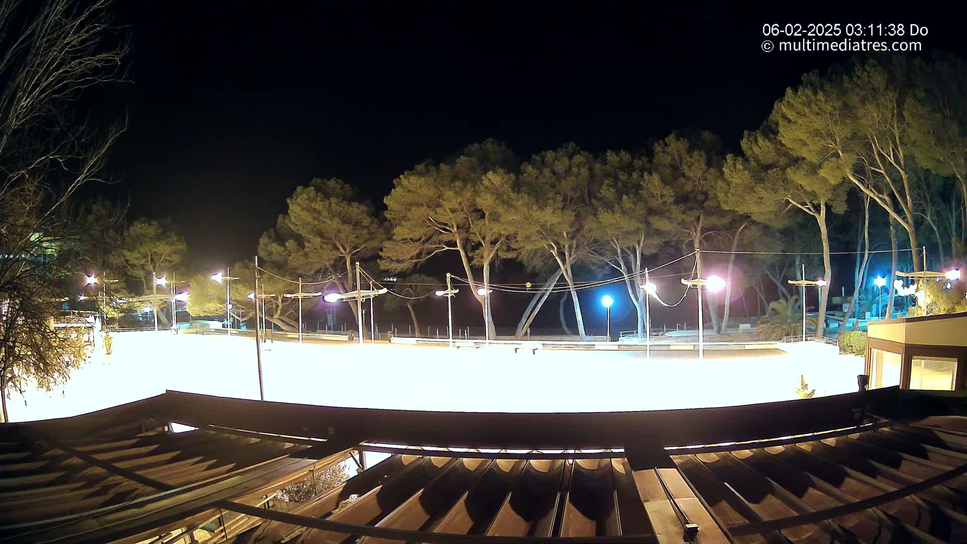 A brightly lit, empty paved area at night, surrounded by tall pine trees and lit by numerous lamps, is viewed from an elevated position partially obscured by a structure’s roof.