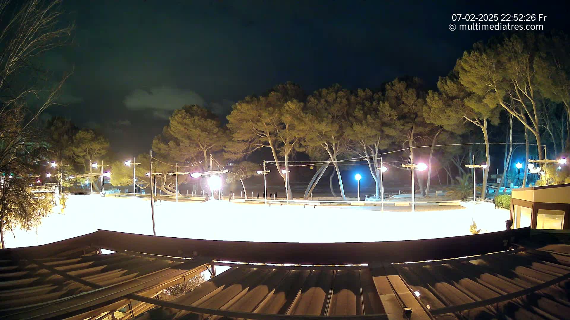 A brightly lit, empty skating rink at night, surrounded by tall trees under a partly cloudy sky.