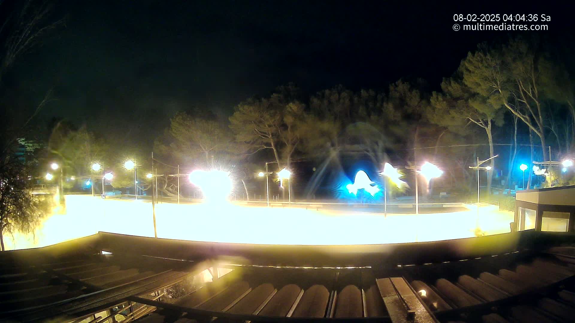 A brightly lit, flat, outdoor area at night, surrounded by trees and streetlights, appears to be experiencing windy conditions.