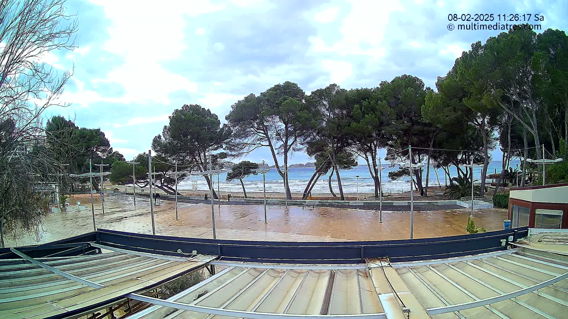 A mostly cloudy day shows a wet paved area next to a beach with several pine trees and a calm sea in the background.