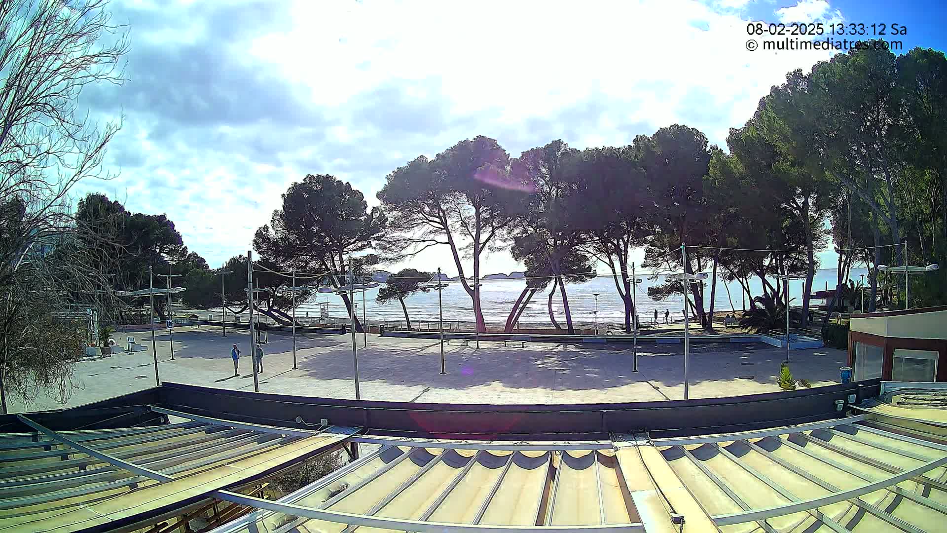 A mostly sunny day with some clouds overhead shows a view of a beach with calm water, several people walking along the shore, and a row of pine trees between the beach and a paved area with a retractable awning.