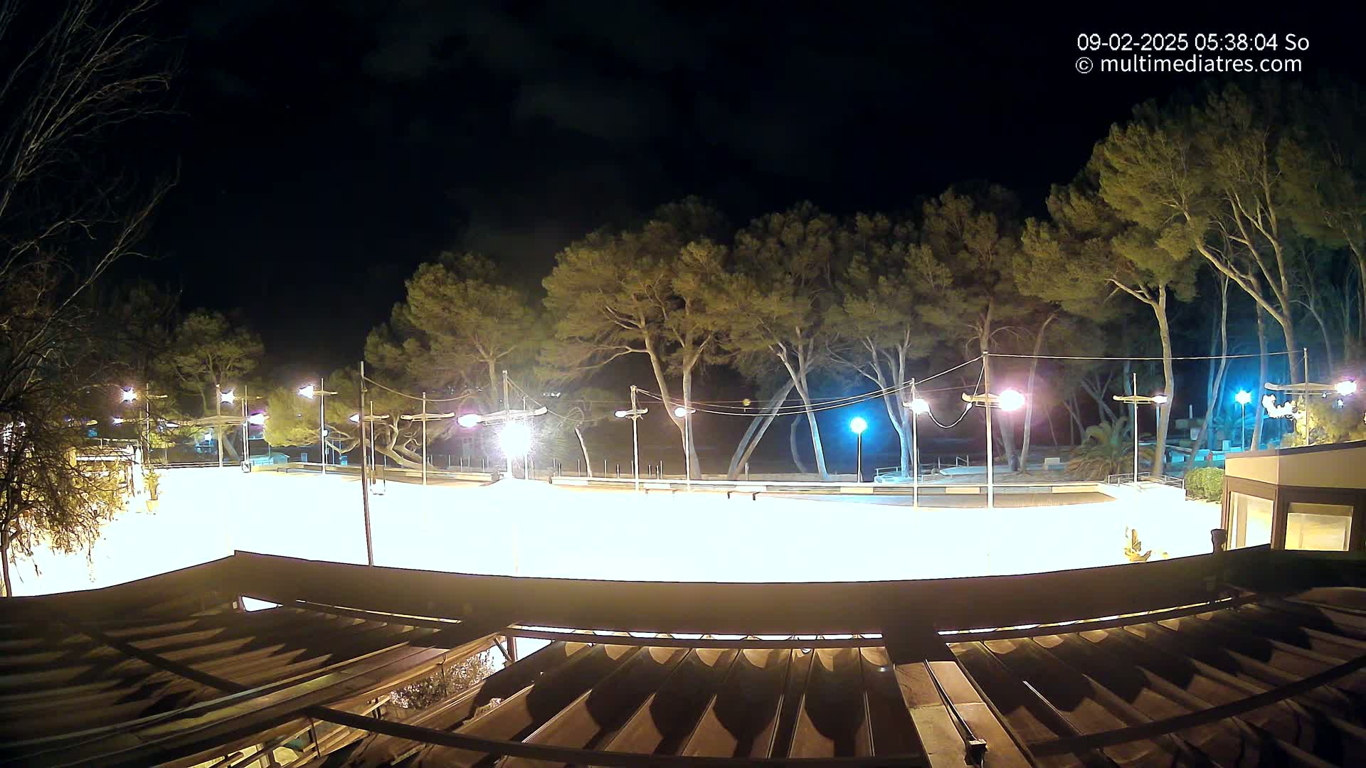 A brightly lit, empty paved area at night, surrounded by tall trees under a dark, cloudy sky.