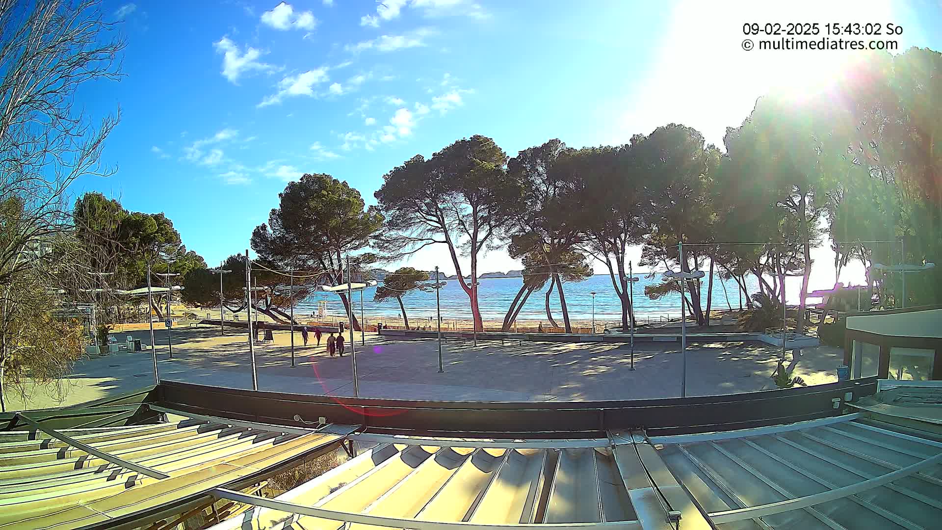 A sunny day reveals a beach with calm water, lined with pine trees, and an empty plaza in the foreground, partially obscured by a retractable roof structure.