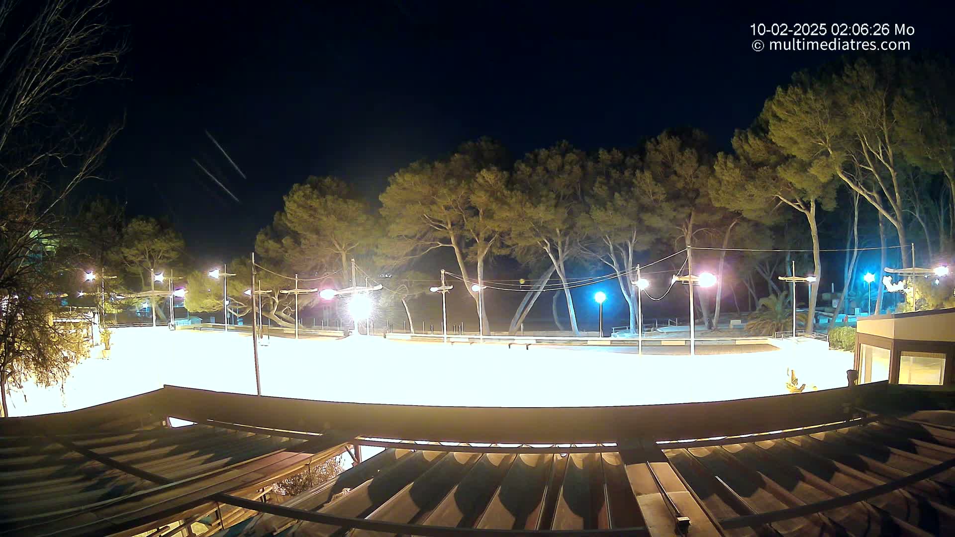A brightly lit, empty plaza or rink at night, surrounded by tall trees under a clear, dark sky with faint light streaks.
