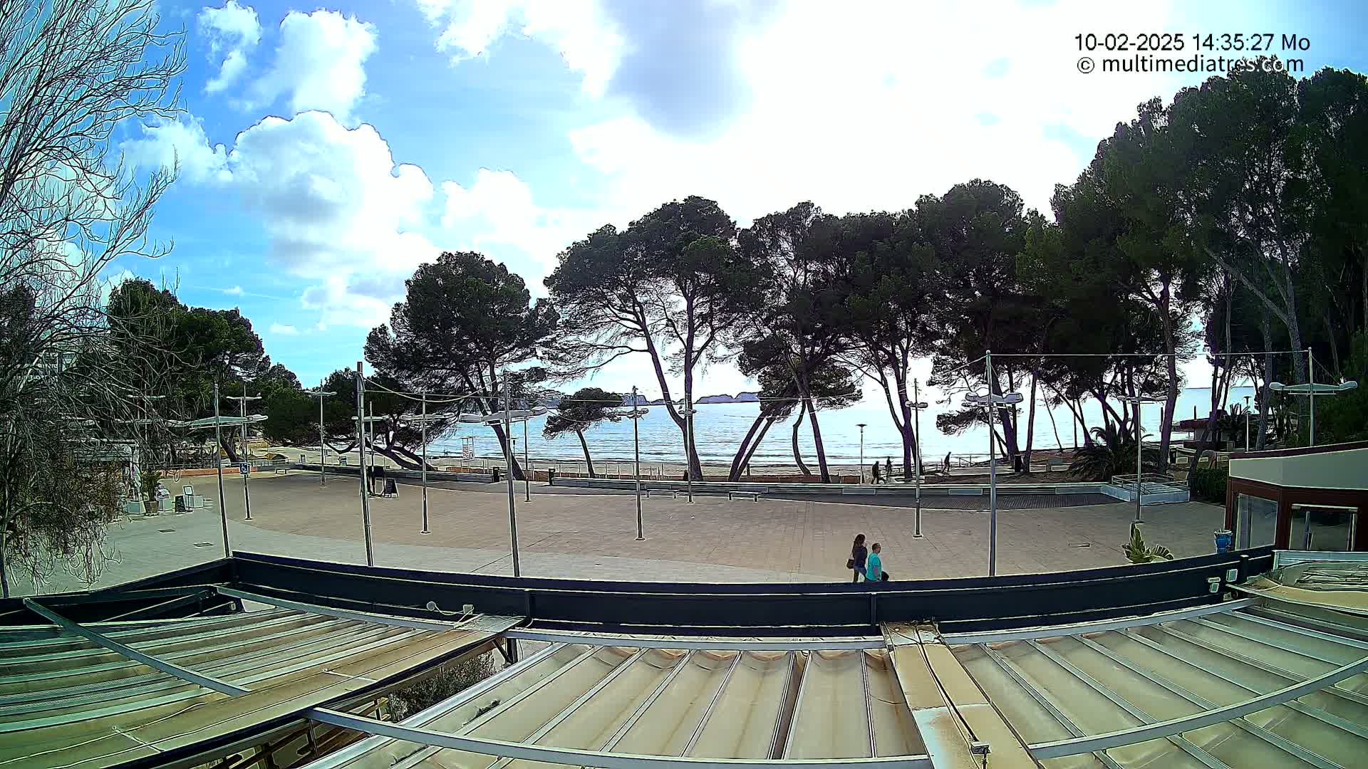 A partly sunny outdoor scene shows a paved plaza with a few people walking, lined with pine trees overlooking a calm, light-blue ocean beach.