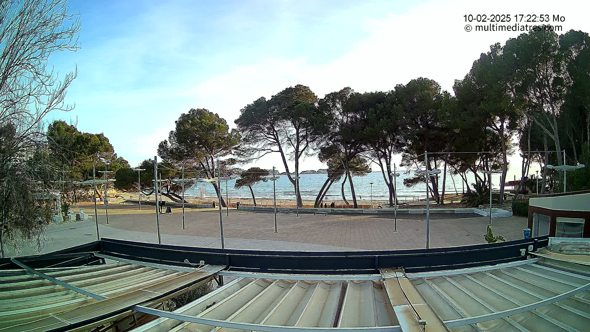 A mostly sunny outdoor scene shows a paved plaza overlooking a sandy beach and calm ocean, with a row of pine trees between the plaza and the water.