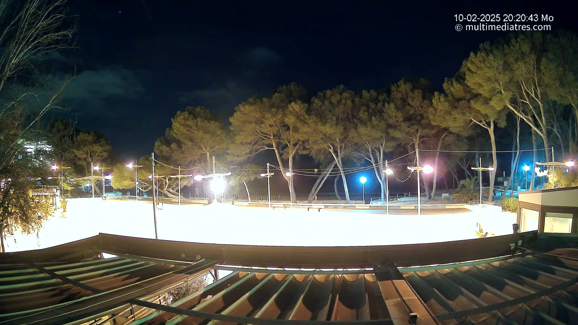 A brightly lit, empty plaza at night, surrounded by tall trees under a partly cloudy sky.