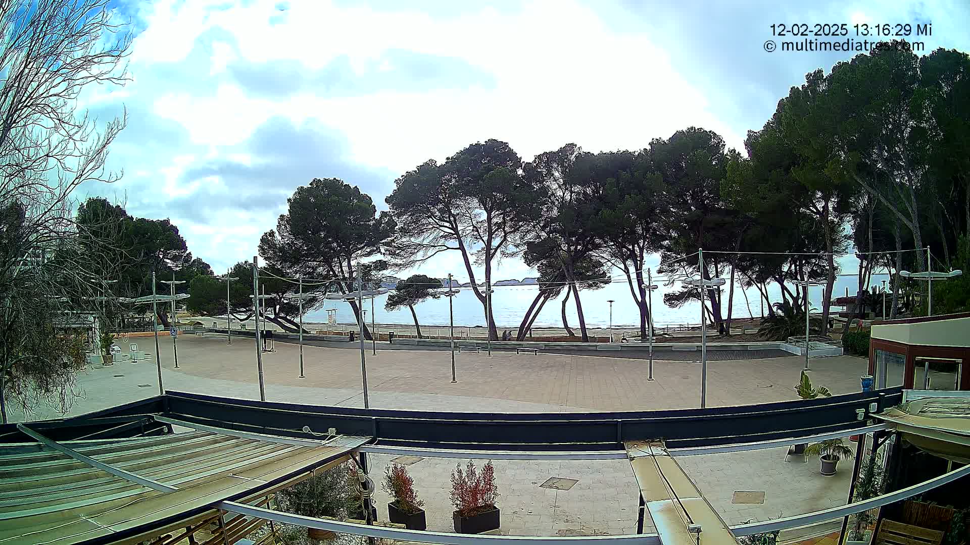 A mostly sunny day overlooks a paved plaza, lined with trees, leading to a calm ocean beach.
