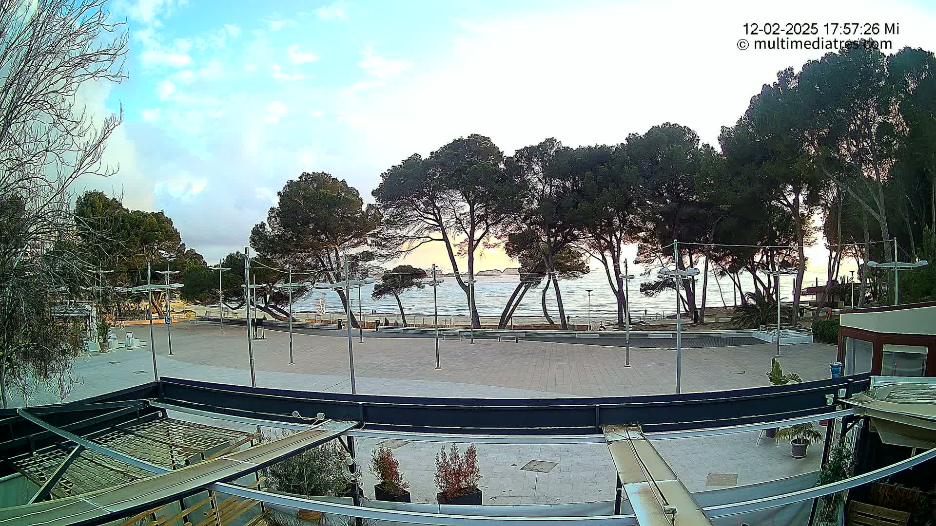A paved plaza overlooks a calm ocean beach at sunset, lined with tall, dark green trees under a partly cloudy sky.