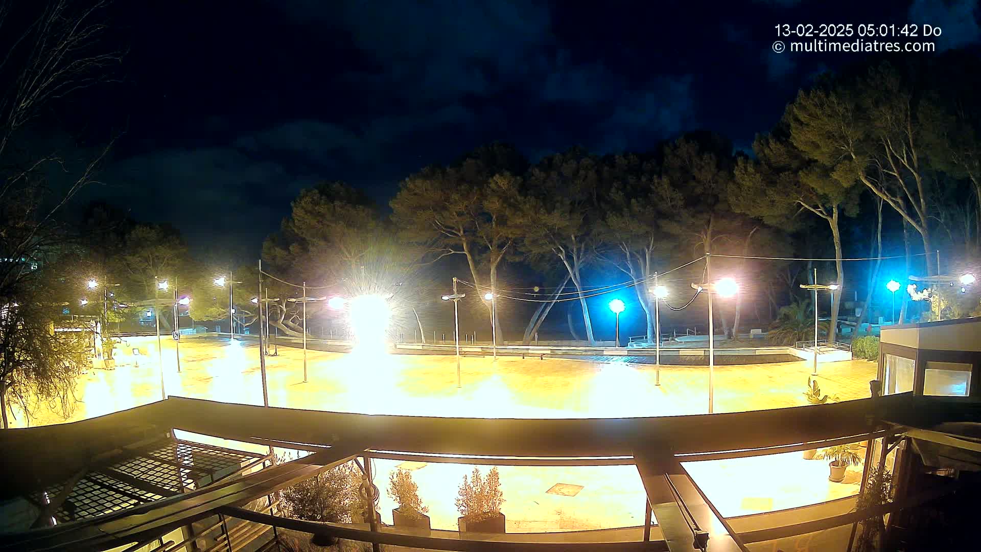 A brightly lit, empty plaza at night under a partly cloudy sky, surrounded by trees and buildings.