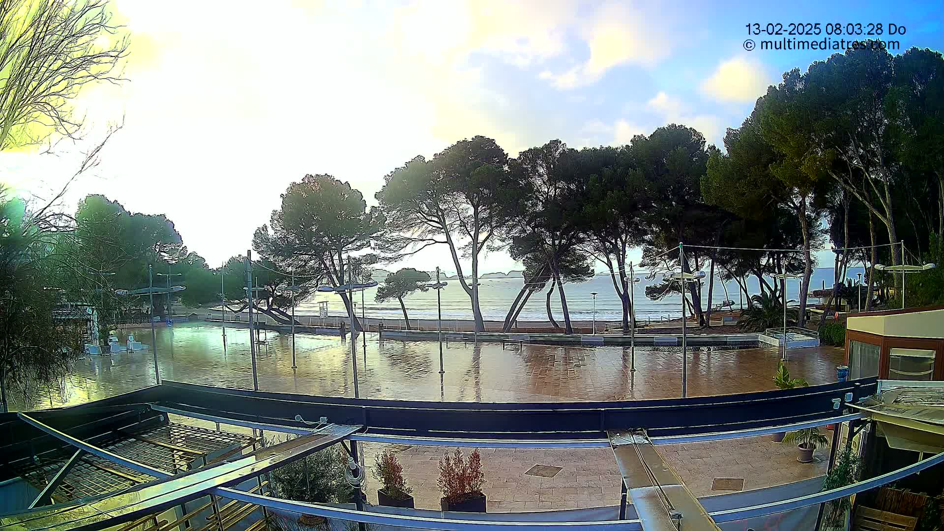 A flooded plaza next to a beach with calm waves under a partly cloudy sky.