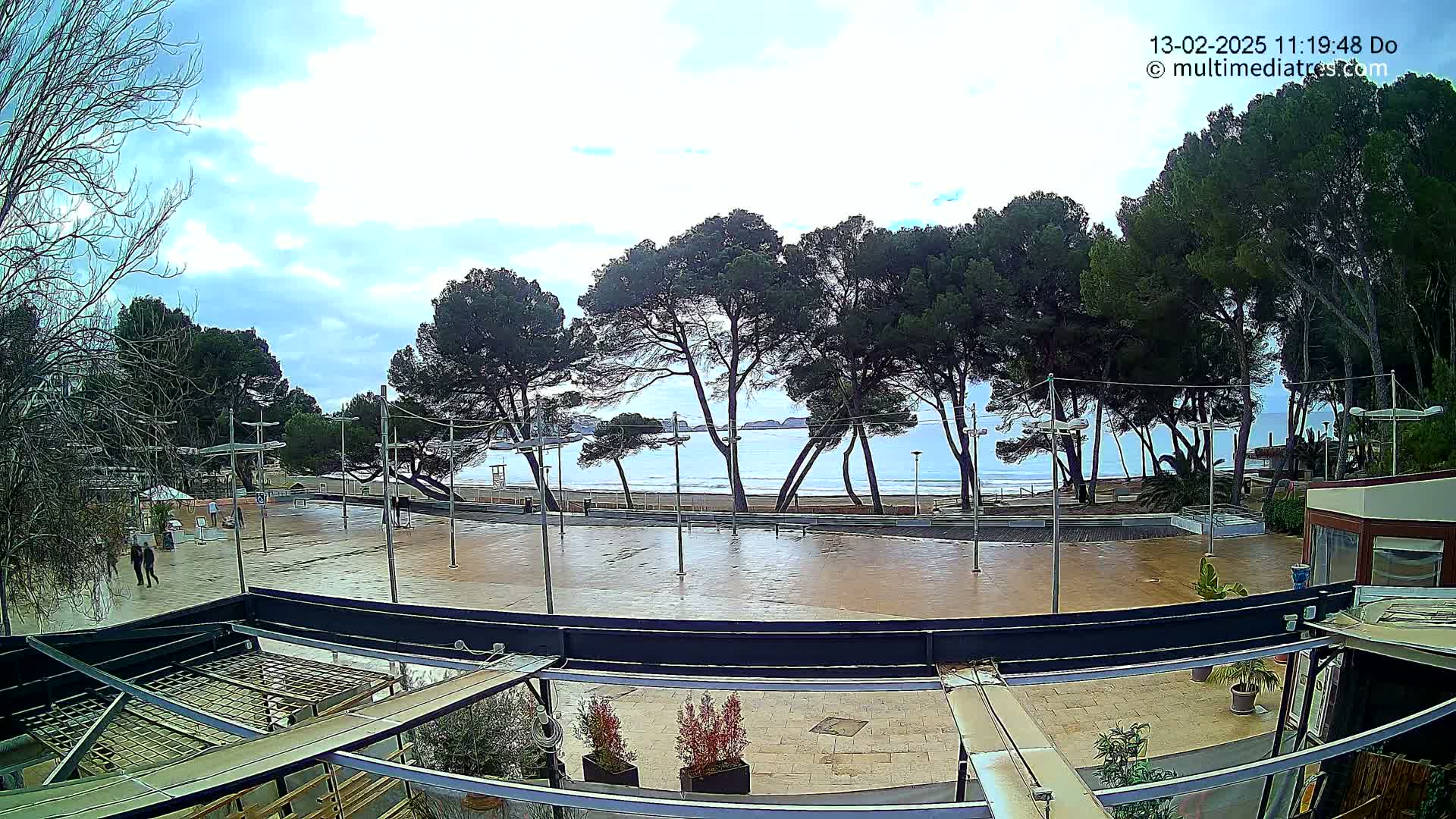A wet plaza overlooks a calm ocean beach lined with pine trees under a partly cloudy sky.