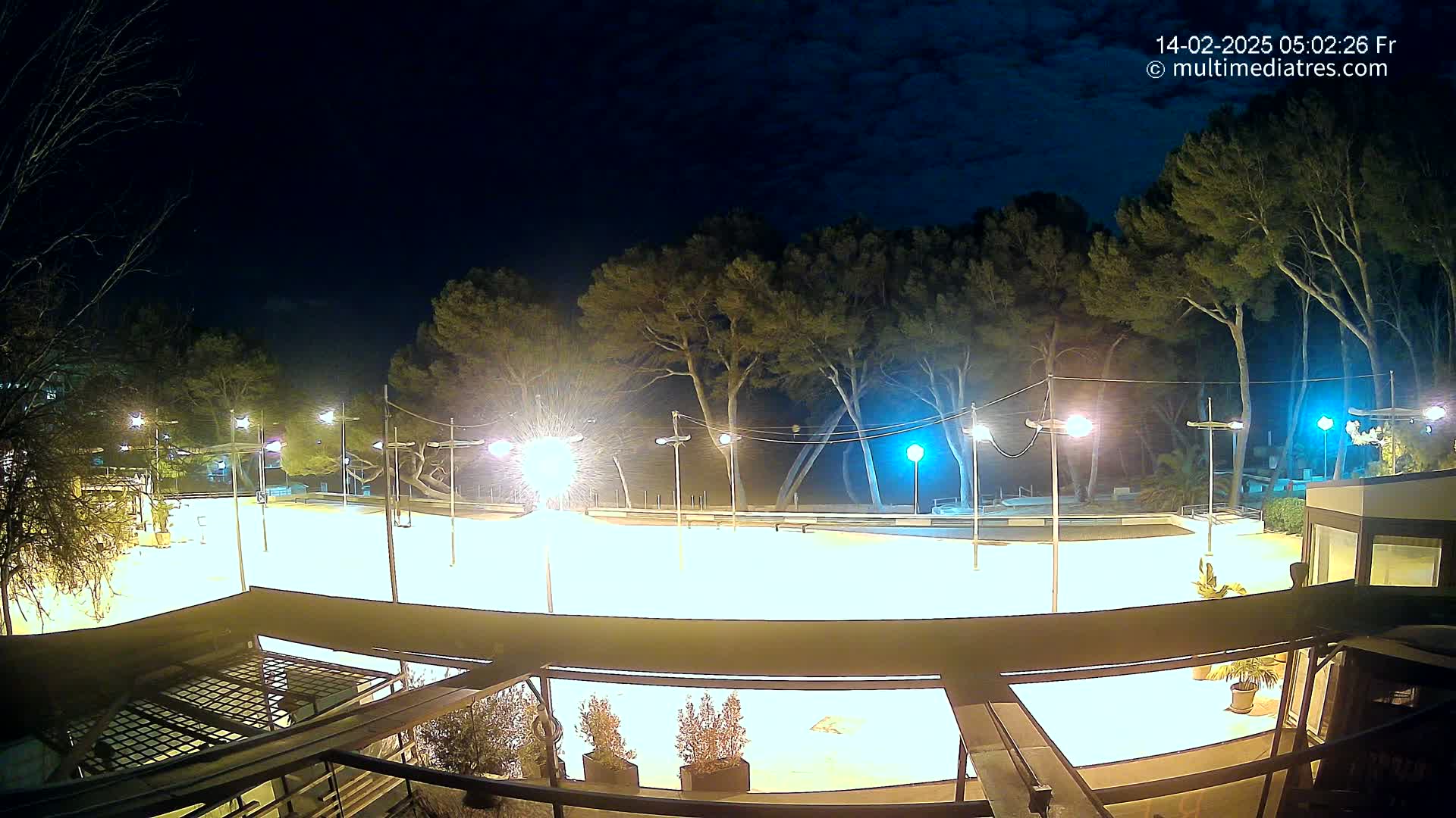 A brightly lit, empty outdoor rink is surrounded by trees under a partly cloudy night sky.