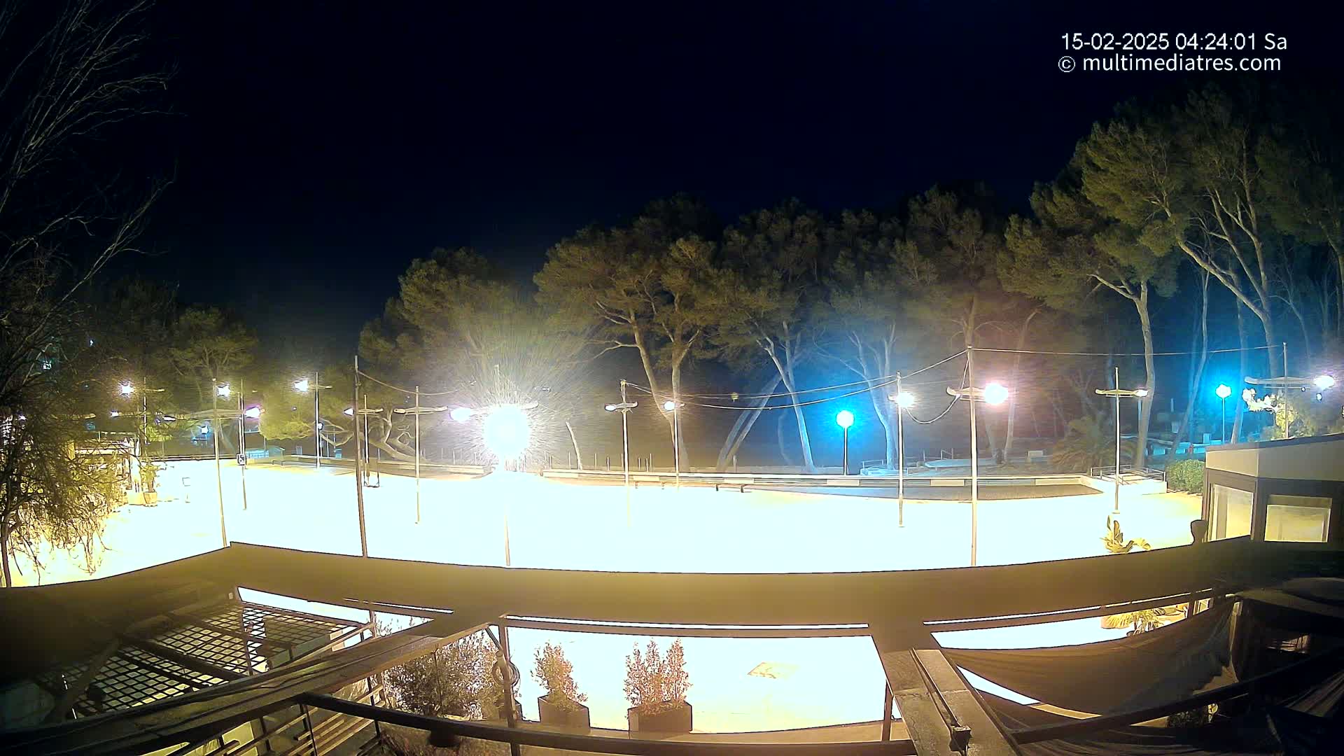 A brightly lit, empty outdoor skating rink at night, surrounded by trees and illuminated by numerous lampposts under a clear, dark sky.